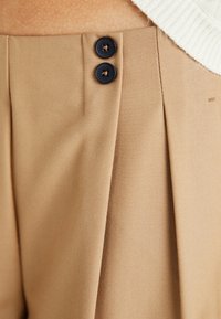 Pantalons plissés beige en tissu texturé, avec deux boutons foncés à la taille et une finition lisse.