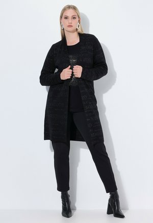 Cárdigan de punto negro con un frente abierto, que presenta letras texturizadas. Combinado con una blusa negra y pantalones ajustados, completado con botas de tacón negras.