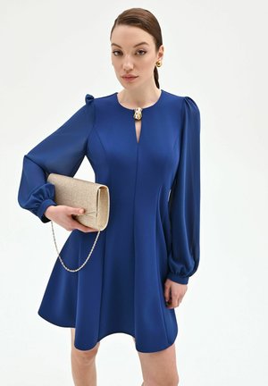 Femme portant une robe bleue à manches longues avec une boucle dorée, tenant une pochette dorée scintillante avec une bandoulière chaîne.