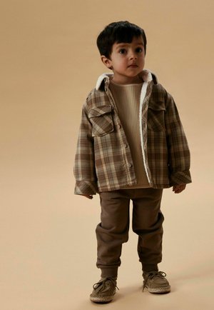 Malé dítě stojí oblečené v hnědém kostkovaném kabátu s bílou fleecovou podšívkou, béžovém svetru, hnědých kalhotách a hnědých botách na béžovém pozadí.
