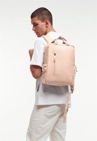 Béžový batoh s obdélníkovým designem, vyrobený z měkkého materiálu, s přední kapsou na zip a horním uchycením, nošený na jednom rameni.