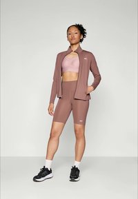 Brun atletisk outfit bestående af en tætsiddende jakke, sports-bh og shorts. Funktioner inkluderer en glat tekstur og minimalistisk design med hvide accenter.