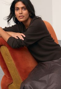 Brown sweater and dark leather skirt. Woman leans on an orange velvet chair with wooden accents. Sleek, textured fabric contrasts with smooth wood.