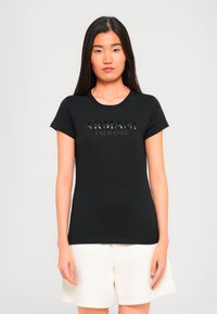 Kvinne som står iført en svart Armani Exchange t-skjorte og hvite shorts mot en enkel lys bakgrunn.