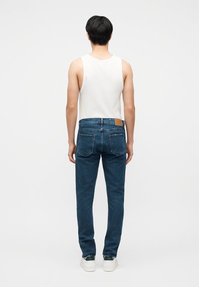 Mężczyzna stoi tyłem, ubrany w białą bezrękawnik, niebieskie dżinsy i białe trampki, na tle jasnego, jednolitego tła.