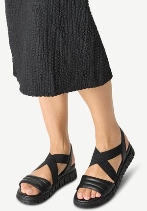 Schwarze Sandalen mit breiten Kreuzriemen, einem Obermaterial aus strukturiertem Stoff und einer dicken, gerillten Sohle. Offene Zehenpartie, die die Zehen zur Schau stellt.