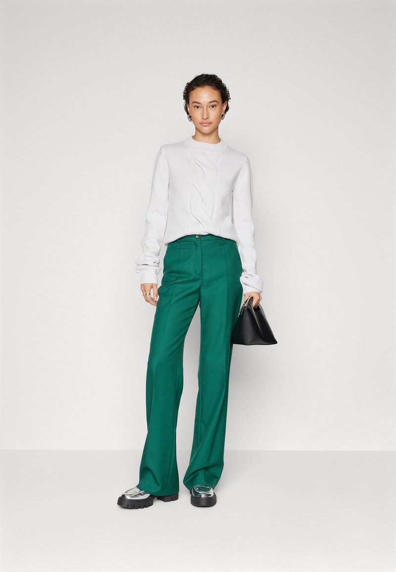 Donna che indossa un maglione bianco a coste, pantaloni larghi verdi a vita alta, scarpe con zeppa argentate e tiene in mano una piccola borsa nera.