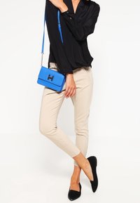 Donna che indossa pantaloni slim beige, blusa nera, ballerine nere e tiene in mano una borsa a tracolla blu brillante con tracolla a catena.