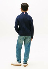 Jūras spēku krāsas adītā džemperis ar augstu apkakli un sarkanām un baltām akcents. Saskaņots ar gaiši ziliem džinsa biksēm un baltām sporta apaviem.