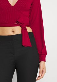 Top de manga longa vermelho com textura de cordas e decote em V, acompanhado de calças pretas justas; o tecido parece suave e elástico.