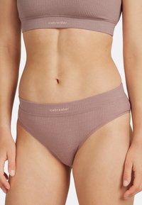 Kobieta w mauve ribrowym staniku sportowym i dopasowanych majtkach o wysokim kroju z logo Icebreaker na pasku, na białym tle.