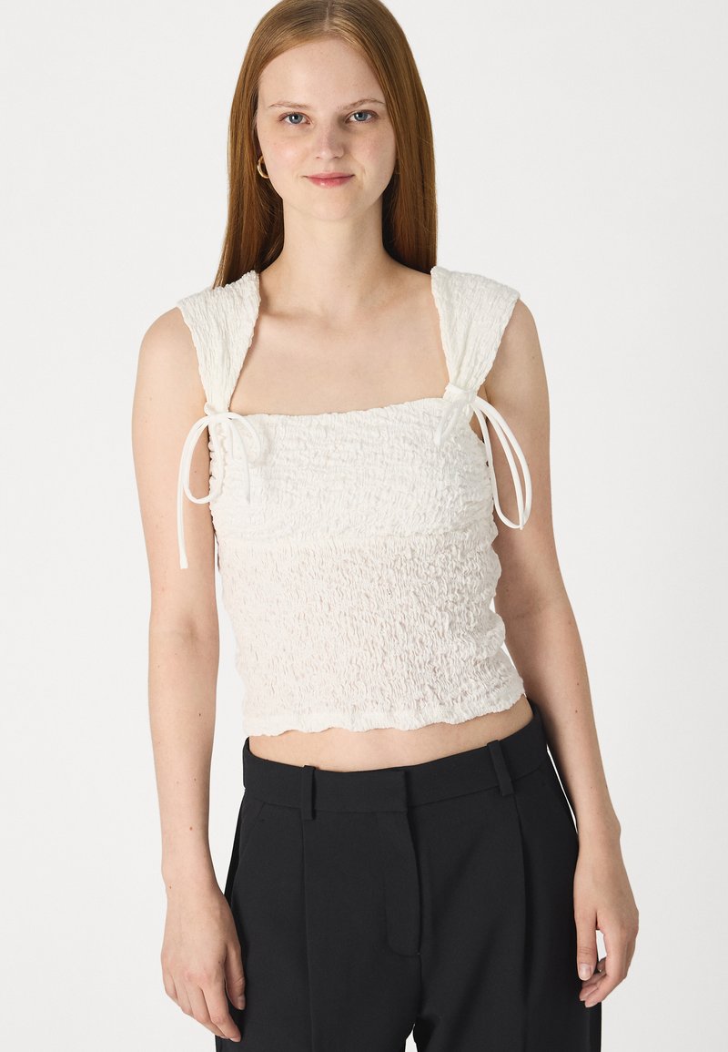 Biała, fakturowana crop top z kwadratowym dekoltem, elastycznym materiałem, szerokimi ramiączkami oraz wiązaniami na ramionach. Noszona z czarnymi, dopasowanymi spodniami.
