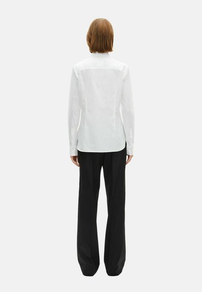 White long-sleeve shirt with a smooth texture and fitted design, paired with loose black trousers. Shirt features subtle back seam detailing.