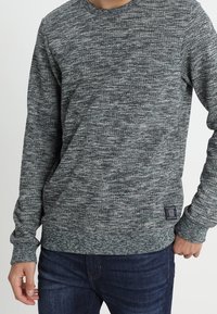 Suéter gris de manga larga con un patrón texturizado, cuello redondo y una pequeña etiqueta de tela en la parte inferior izquierda. Llevado sobre jeans oscuros.