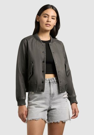 Jonge vrouw die een grijze bomberjack draagt over een zwarte crop top en lichtgrijze gescheurde denim shorts, staande tegen een effen achtergrond.
