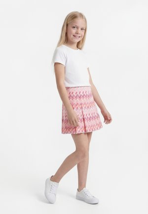 Smilende jente med langt blondt hår står iført en hvit kortermet skjorte, rosa mønstret skjørt og hvite joggesko på en hvit bakgrunn.