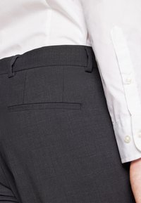 Tamne tamni sivi krojaški hlače od teksturiranog materijala, s zadnjim džepom i petljama za pojas, u kombinaciji s bijelom košuljom s dugim rukavima.
