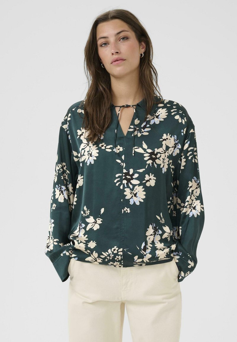 Blusa floreale verde scuro con scollatura a fessura, maniche lunghe e vestibilità comoda. Presenta motivi floreali in crema e azzurro chiaro.