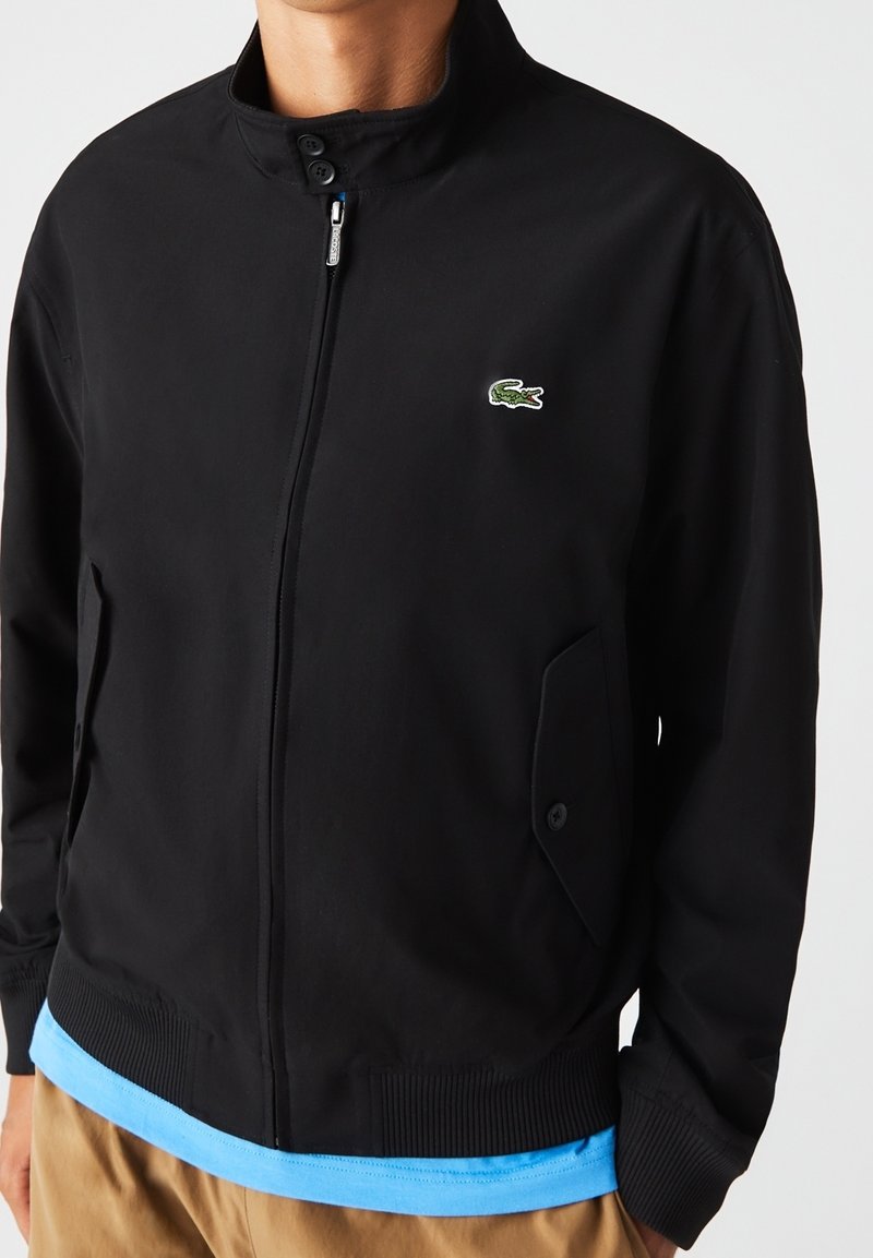 Homme portant une veste noire zippée à col montant avec un logo crocodile vert sur la poitrine, les mains dans les poches.