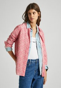 Giacca rosa con motivo floreale e maniche arrotolate, indossata sopra un top bianco, abbinata a jeans blu.