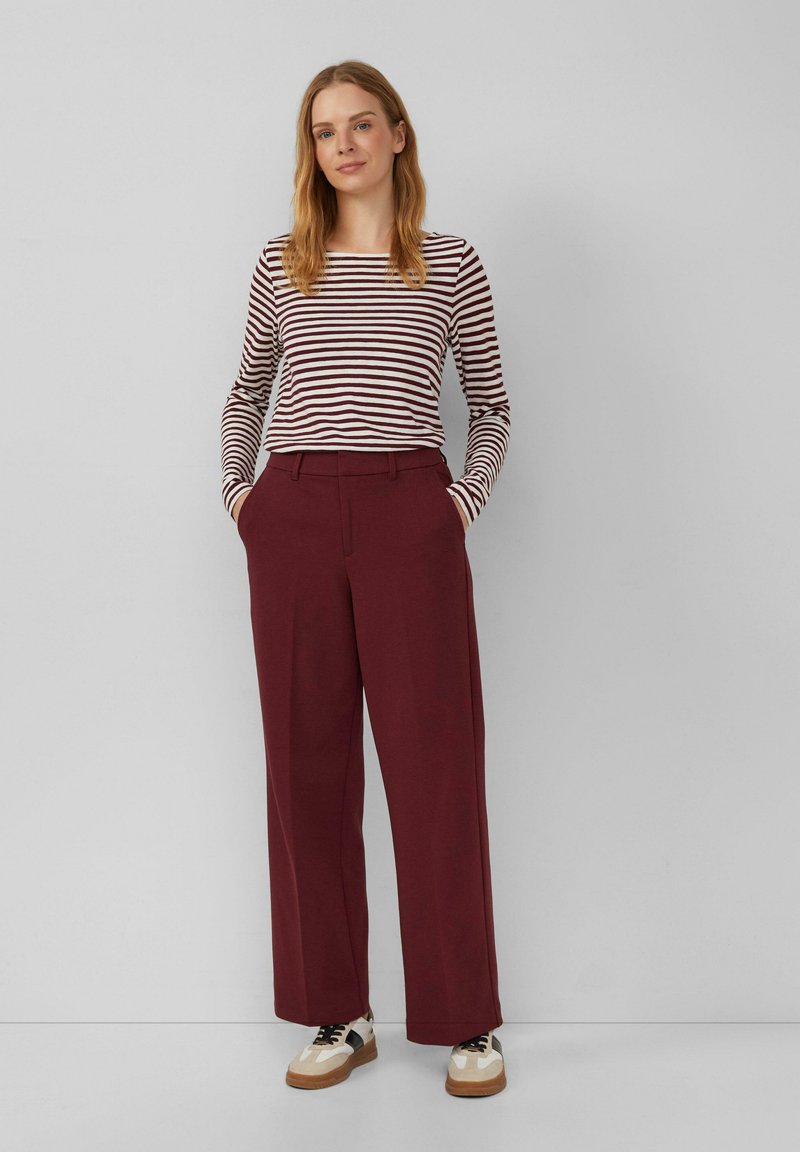 Gestreepte langemouwtop in bordeauxrood en wit, gecombineerd met bordeauxrode brede broek. De schoenen hebben witte en zwarte accenten met een bruine zool.