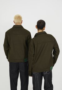 Olivenfargede jakker laget av myk stoff, med krage og design på baksiden. Båret med mørke jeans, som viser kontrasterende teksturer og sømmer.