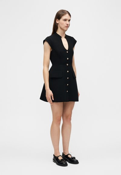 Femme portant une robe courte noire avec des boutons dorés sur le devant et des poches à rabat, associée à des chaussures Mary Jane noires.