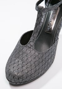 Chaussure à talons hauts gris avec une surface brillante et texturée et des découpes. Présente une bride en T et un bout rond. Doublure intérieure lisse.