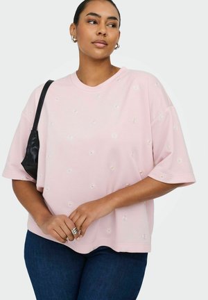 Donna che indossa una maglietta rosa chiaro oversize con piccole margherite bianche, jeans scuri, anelli d'argento, orecchini a cerchio e una borsa a tracolla nera.