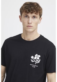 Sort bomulds T-shirt med et hvidt blomstermotiv og teksten "ARCHIVE & SOLID © 1999" på venstre brystområde. Rund halsudskæring.