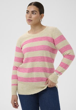 Frau, die einen beige-rosa gestreiften Pullover, blaue Jeans, ein silbernes Armband und Creolen trägt und mit der Hand in der Hüfte vor einem weißen Hintergrund posiert.