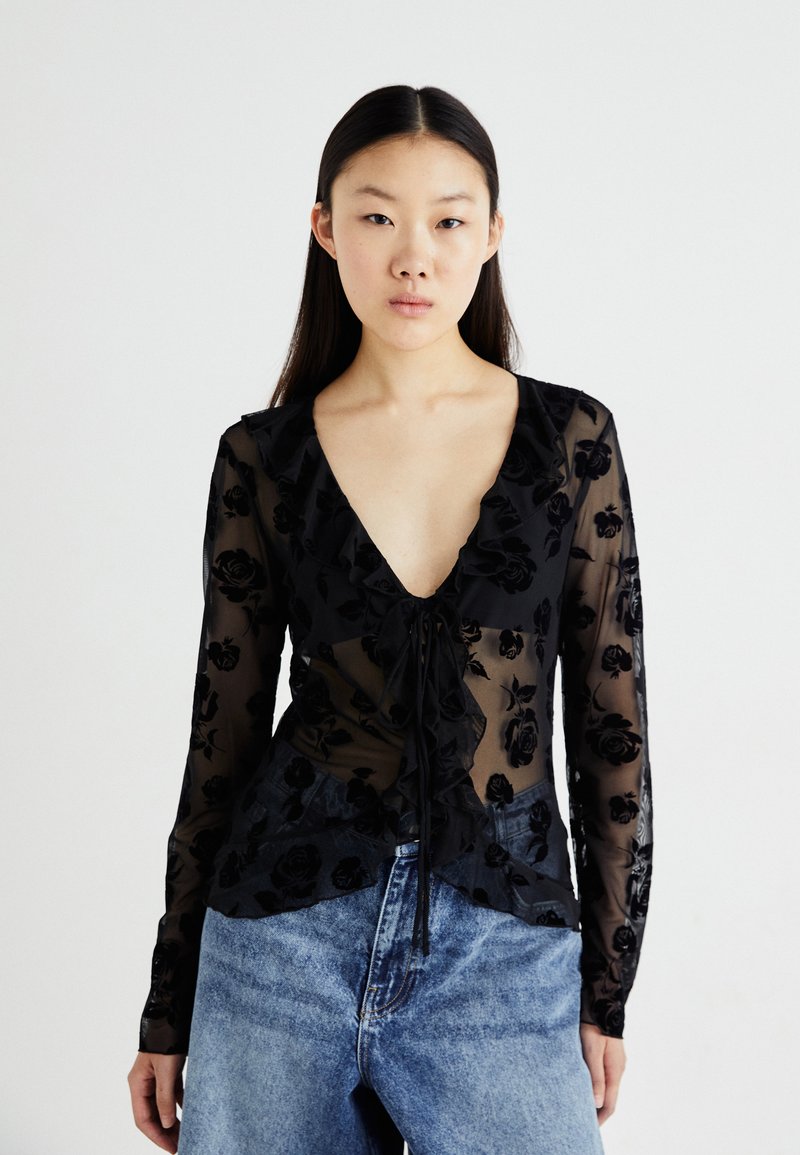 Blusa nera trasparente con motivo floreale, scollatura a V profonda, bordi arricciati e dettaglio con fiocco davanti, abbinata a jeans blu chiaro a vita alta.
