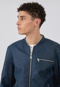 Veste de pilote bleu marine avec une texture lisse, col rond, fermeture éclair frontale et une poche poitrine avec une fermeture éclair métallique.