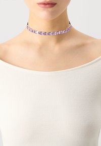 Chokers ar purpursarkanām taisnstūrveida dārgakmeņiem, iestatītiem zelta tonī, kombinēti ar melnu auklu. Fona teksturēta balta blūze bez piedurknēm.