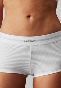 Bijele rebraste pamučne bokserice s mekanim pojasom sivi naglasci i "Calvin Klein" logom u crnoj boji. Glatka tekstura.