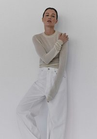 Jasno beżowa bluzka z długim rękawem o prążkowanej strukturze i marszczonymi mankietami, połączona z białymi spodniami o szerokich nogawkach i wysokim stanie. Gładkie tło.