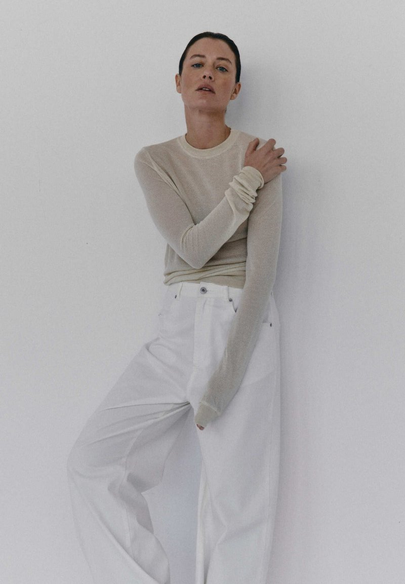 Jasno beżowa bluzka z długim rękawem o prążkowanej strukturze i marszczonymi mankietami, połączona z białymi spodniami o szerokich nogawkach i wysokim stanie. Gładkie tło.