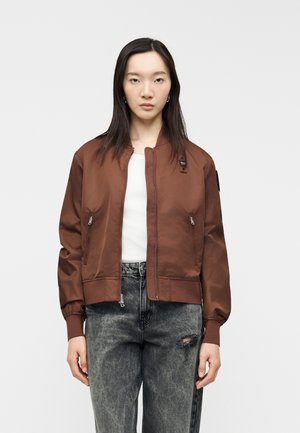 Νέα γυναίκα με καφέ μπουφάν bomber πάνω από άσπρο πουκάμισο και ξεθωριασμένο μαύρο τζιν με σκισίματα, στέκεται μπροστά σε ένα απλό φόντο.