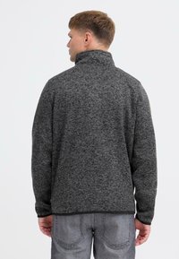 Grå fleecejakke med høj krave, tekstureret overflade og lange ærmer. Matcher med grå jeans, set fra ryggen.