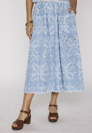 Femme portant une jupe midi bleu clair avec broderies florales ajourées blanches, des sandales marron à talons et des bracelets dorés à une main.