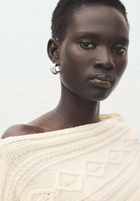 Strikket cremefarvet sweater med teksturerede mønstre, off-shoulder design og ribkantdetaljer. Sølv øreringe i ringform fuldender looket.