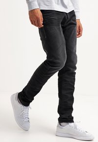 Person wearing black slim jeans and white sneakers, standing with one leg crossed over the other on a white background.