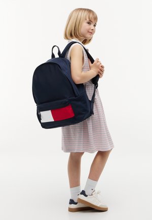 Niña joven con vestido de rayas, lleva zapatillas blancas y una mochila grande azul marino con bolsillo delantero rojo y blanco.