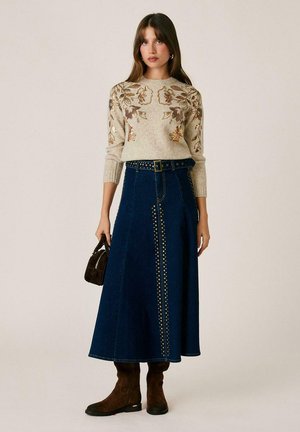 Mujer con suéter beige con flores, falda midi azul oscuro con tachuelas y cinturón, botas marrones, sosteniendo un bolso pequeño marrón, de pie frente a un fondo liso.