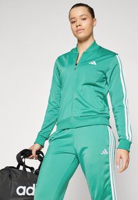 Grön atletisk träningsoverall med vita ränder och logotyp, som har en zip-up jacka och matchande byxor. Matt yta med en åtsittande design.