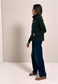Veste zippée vert foncé avec col montant, associée à un jean bleu à jambes larges et des baskets marron. Tissu lisse sans motifs visibles.