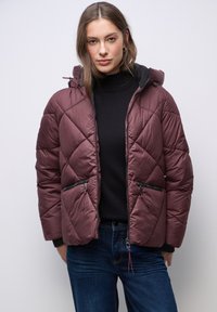 Weinrot gesteppte Pufferjacke mit Kapuze, ausgestattet mit Reißverschlusstaschen an den Seiten, gerippten Bündchen und einer glatten Oberfläche. Getragen über einem schwarzen Pullover.
