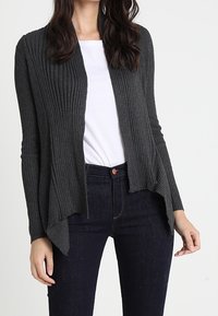 Mörkgrå ribbad cardigan med öppen framkant, långa ärmar och asymmetrisk fåll. Bärs över en vit skjorta och mörk denim.