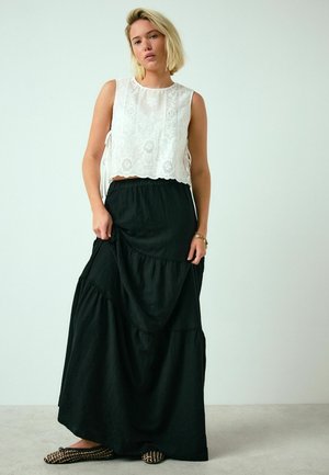 Junge Frau mit kurzen blonden Haaren trägt ein ärmelloses weißes Spitzenoberteil, langen schwarzen gestuften Rock und gemusterte flache Schuhe, steht vor einfachem Hintergrund.