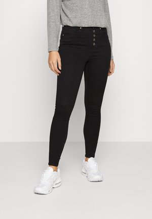 Personne portant un jean skinny noir taille haute avec fermeture à boutons et des baskets blanches épaisses, montrée de la taille aux pieds.